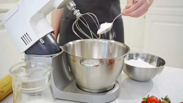 A young woman cook adds a spoonful of flour to prepare dough in a mixer bowl.
