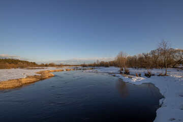 March sunny evening by the river. Blue sky over the horizon. A picturesque landscape, early spring, a river with snow-covered banks, dry grass and bushes. The first thaws, the snow is melting