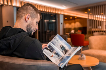 Man reading magazine while sitting in waiting room. Young male looking through travel catalog. He...