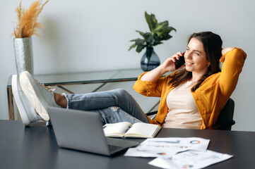 Positive successful caucasian woman, office worker, creative manager, sits in a chair in a relaxed pose with her feet on the table, talking a mobile phone during a work break, looks away, smiles