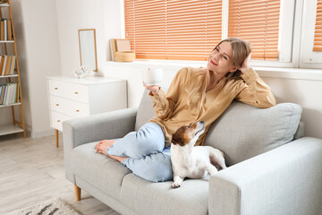 Young woman with cup of tea and Jack Russel terrier sitting on grey sofa at home - obrazy, fototapety, plakaty