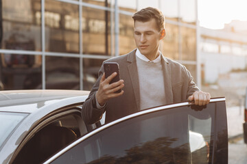 Handsome modern young man with phone entering his car while standing outdoors