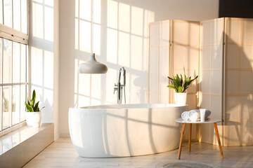 Interior of stylish bathroom with bathtub, table and folding screen near white wall