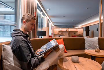 Man reading travel magazine while sitting in lobby. Young male is looking through catalog while...