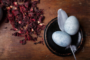 Beautiful diy blue painted easter eggs with dry Hibiscus tea and silver decorative feathers on wooden background, top view