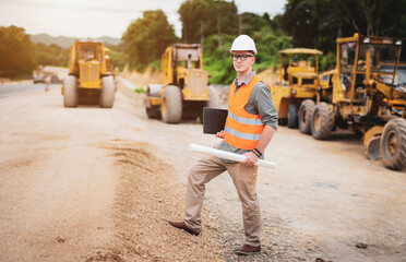Caucasian handsome engineer wear protective helmet stand holding a laptop and roll of paper on road construction site with heavy equipment on the background