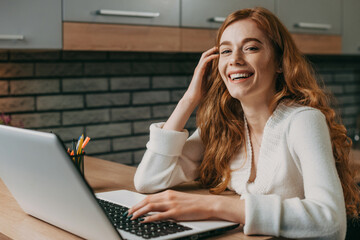Obraz premium Photo of a redheaded positive business woman sitting indoors using laptop computer. Business woman. Business technology.