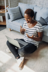 Full body photo of african american woman smiling watching seminar on laptop sitting on the floor at home. Online communication. Business communication