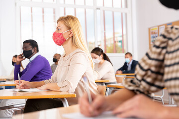 Fototapeta premium Portrait of focused female in protective face mask sitting at desk studying in classroom with colleagues