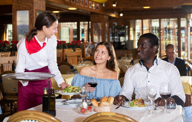 Polite waitress serving food for positive interracial couple at restaurant, putting plate with salad on table