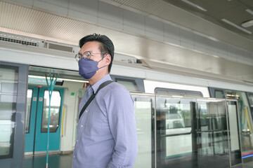 Asian man commuter with face mask on standing on subway train platform. Masked transit concept.