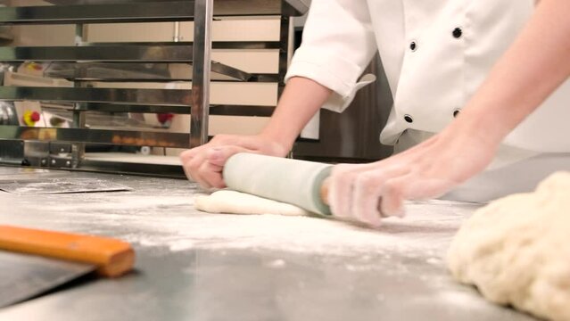 Close-up, chef in white cook uniform with apron sprinkle flour, knead pastry dough with roller, prepare bread, pies, and fresh bakery food, baking in oven at stainless steel kitchen of restaurant.