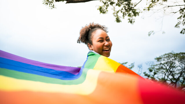 Happy young African woman carrying LGBT rainbow flag symbol and smiling in camera in green city park. Concept of homophobia,diversity,equity, peace and love,freedom,liberty,Lesbian,Feminist,Banner.