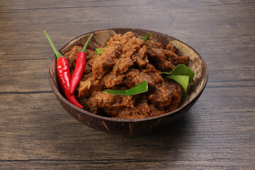 Traditional cooked dry curry beef meat rendang closeup in coconut shell bowl on rustic wooden background - obrazy, fototapety, plakaty