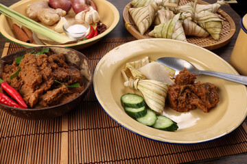 Traditional dry beef curry rendang in coconut shell diamond shaped woven palm leaf pouch steamed glutenous rice cake ketupat kupat tipat on bamboo tray mat ingredients rustic wooden table