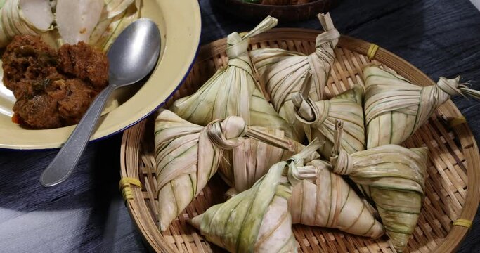 Traditional dry beef curry rendang in coconut shell diamond shaped woven palm leaf pouch steamed glutenous rice cake ketupat kupat tipat on bamboo tray mat ingredients rustic wooden table rotate
