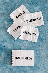 Happiness text on notepad surrounded by Fear Anxiety Stress and Burnout words on torn scrunched up pages, psychology and overcoming your past obstacles