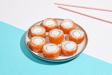 Philadelphia maki roll on ceramic plate with chopsticks. Salmon sushi roll with philadelphia cheese on coloured background. Japanese menu concept. Salmon sushi roll in modern style.
