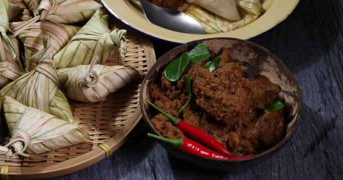 Traditional dry beef curry rendang in coconut shell diamond shaped woven palm leaf pouch steamed glutenous rice cake ketupat kupat tipat on bamboo tray mat ingredients rustic wooden table rotate