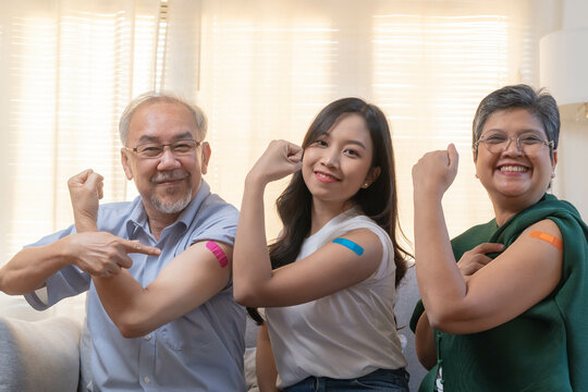 Coronavirus Vaccination, happy asian elderly family with daughter smile strong together, showing bandage on arm protect of covid-19 after injection of vaccine, sitting on couch in living room at home. © KMPZZZ