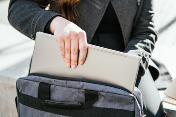 Crop anonymous female freelancer in outerwear sitting on concrete bench and taking netbook from...