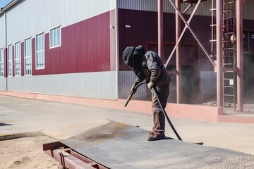 A worker in a special suit is sandblasting metal at an industrial site.
