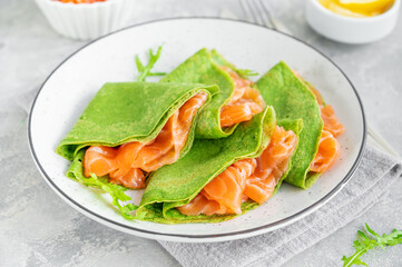 Green spinach crepes or pancakes with smoked salmon and soft cheese on a plate on the gray background. Copy space.