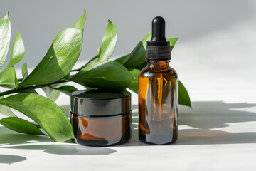 close up of collagen cream in glass jar, serum in amber glass bottle and ruskus branch on gray...