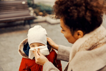 A woman blowing the nose of a little boy at the park in cold weather.