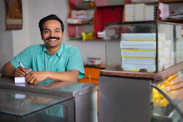 Portrait of Sweet shop owner looking at camera with smile while maintaining accounts at counter
