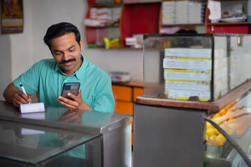 Portrait of Sweet shop owner maintaining accounts by using Smartphone at counter