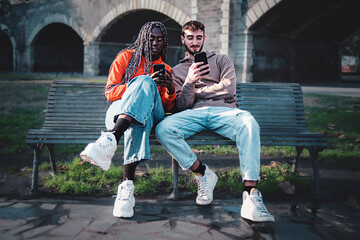 Couple fo young trendy biracial young people using smartphone sitting together on a bench in the public city park - young friends using social network and the internet - technology addiction concept
