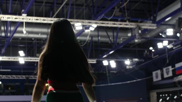 Long haired lady cheerleader shakes pompoms to support players at hockey tournament on modern ice arena backside view