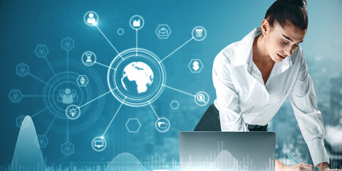 Attractive european businesswoman working at desktop with laptop and abstract glowing business data and earth hologram on blurry blue city background. Communication and media concept. Double exposure.