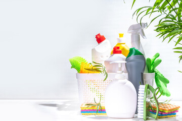 Fototapeta na wymiar Summer cleaning concept. Plastic basket with different detergents and cleaning supplies bottles, accessories for cleaning house with palm leaves on a light white background