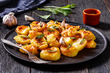 crispy roasted squashed potatoes with garlic and sage leaves on black plate on dark wooden table with ingredients at background, horizontal view from above