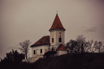 An eerie chapel at the top of the hill. The sky is threatening, the trees are very dark, the chapel is scary, the roofs are dirty red. The atmosphere is very scary stressful.