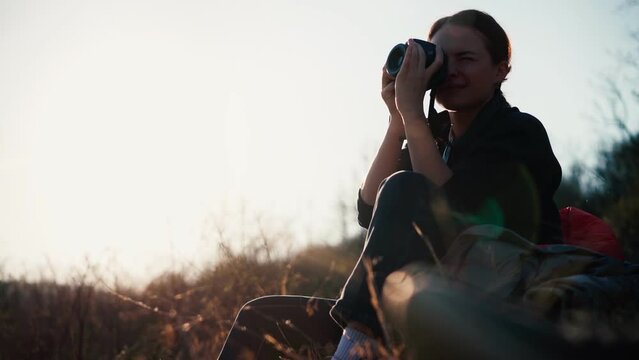 A young beautiful woman traveler taking a picture with her camera and enjoying the view while sitting on the hill at sunset time.