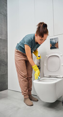 A woman in yellow rubber gloves wipes the toilet bowl with a rag. Toilet cleaning and disinfection.