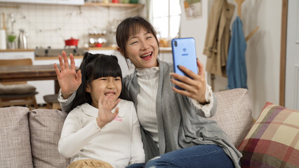 cheerful asian mother and daughter having video chat with friends at home. they smile and wave hands to cellphone together on the couch