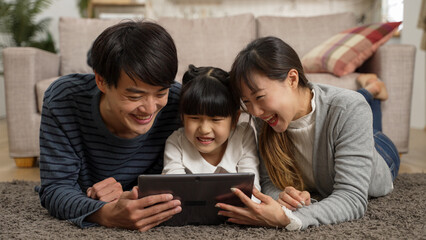 Happy Asian girl lying on floor with her father and mother and having fun with online educational games on touchpad. Cheerful parents point at screen while giving advice to daughter