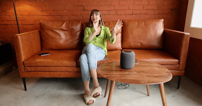 Young woman using phone and controlling home devices with a voice command while sitting relaxed on leather couch. Concept of smart home and leisure time with gadgets