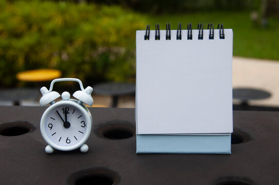 Alarm clock pointing at 11 o'clock with blank notepad. Blurred park background. Copy space concept