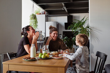 Happy lesbian couple laughing during breakfast at table while their little son talking funny jokes - obrazy, fototapety, plakaty