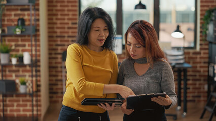 Asian women comparing statistics on clipboard and tablet, planning business strategy to grow sales and e commerce management. Colleagues meeting to brainstorm new ideas in startup office.