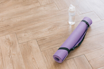 Rolled purple yoga mat and clear plastic water bottle on a wooden surface with natural light. Gender-neutral fitness yoga and exercise props at home or gym banner with copy space. Active lifestyle. - obrazy, fototapety, plakaty