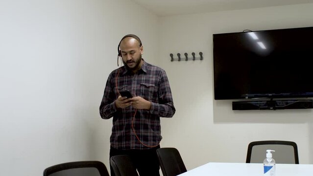 An Indian man pacing up and down a boardroom talking to a colleague on his mobile phone, the man focused on his conversation speaking into a microphone headset connected to his phone