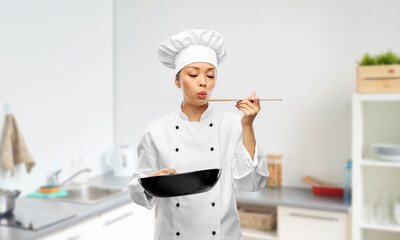 cooking, culinary and people concept - female chef with frying pan blowing to spatula and tasting food over kitchen background