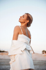 Fototapeta na wymiar Gorgeous lady in stylish white suit standing on sand