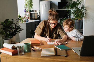 Young mother showing how to write to her son while they doing homework together at table in kitchen
